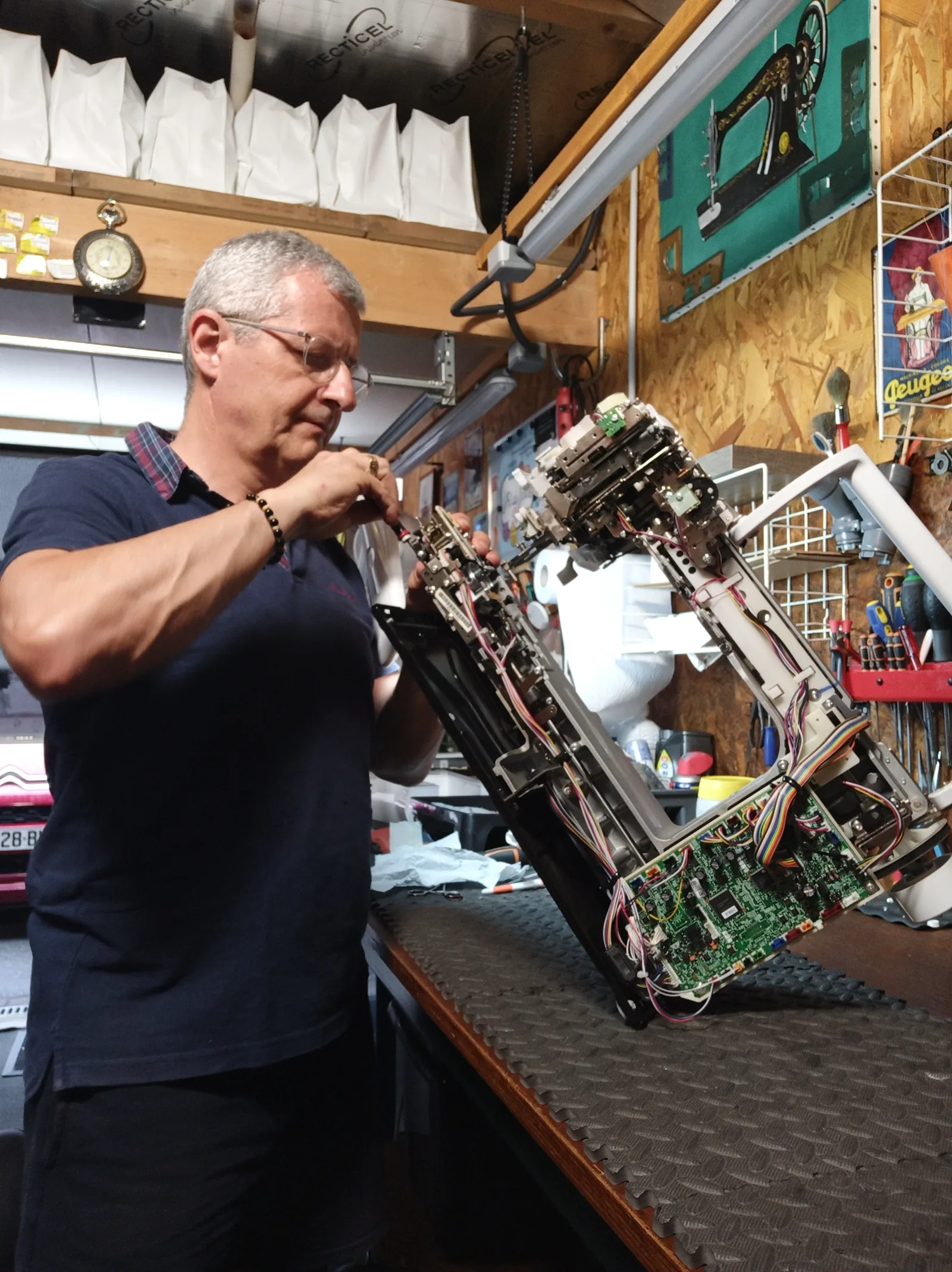Un réparateur ajuste au tournevis le mécanisme électronique d'une machine à coudre démontée, sur son établi.